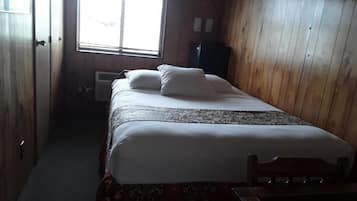 A small, dimly lit room with a single bed, wooden walls, and a window with blinds.