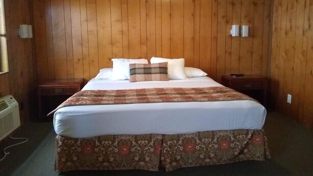 A room with a wooden paneled wall, a bed with a floral bedspread, two bedside tables, and a wall-mounted heater.