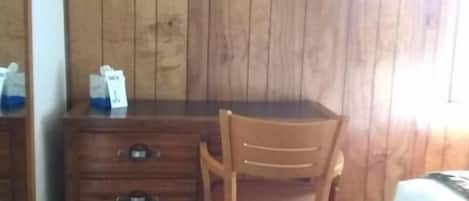 Un aparador de madera con cajones, una silla de madera, una cama con colcha roja y una pared de madera.