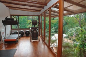 A gym with exercise equipment, a treadmill, and large windows overlooking a garden. - Sunrise Studio at Taru Rentals Ocean Views (Montezuma)