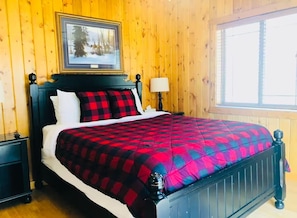 A wooden-paneled room with a bed, a nightstand, a lamp, and a framed picture on the wall.