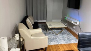 Un salon avec un canapé, une table basse, un téléviseur fixé au mur et une fenêtre avec des rideaux.