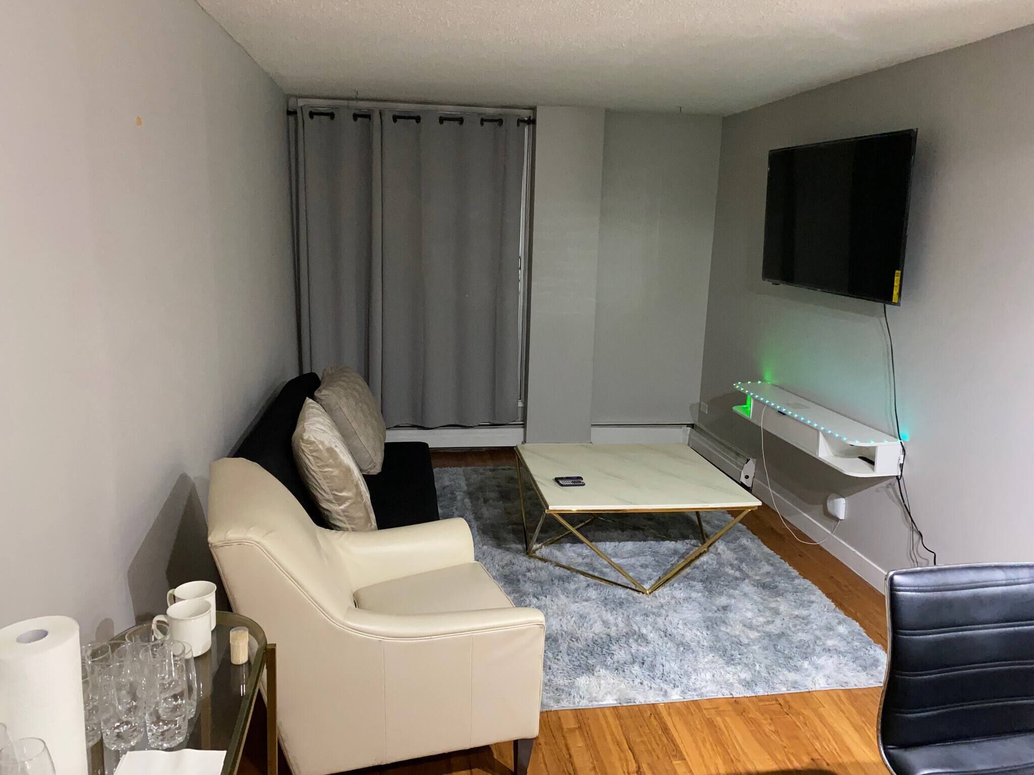 Une petite pièce moderne, avec un canapé en cuir blanc, une petite table sur laquelle se trouve un smartphone, un téléviseur fixé au mur et un rideau gris.