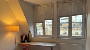 Un dormitorio con cama, escritorio, silla, lámpara y vistas a edificios a través de las ventanas.
