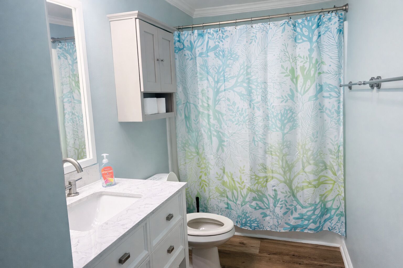 Une salle de bain comprenant des toilettes, un lavabo avec un plan de travail en marbre, un meuble au-dessus du lavabo et un rideau de douche orné d’un motif de feuilles.