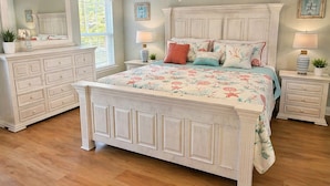 A bedroom with a large bed, two bedside tables, a dresser, a ceiling fan, and a window with blinds.
