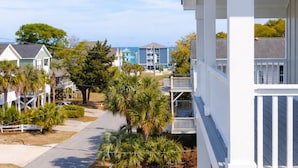 A residential area with a white building featuring a balcony, a street with palm trees, and other houses in the background.