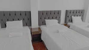 Een hotelkamer met drie bedden, elk voorzien van een grijs gestoffeerd hoofdeinde, witte beddengoed en een houten nachtkastje ertussen.