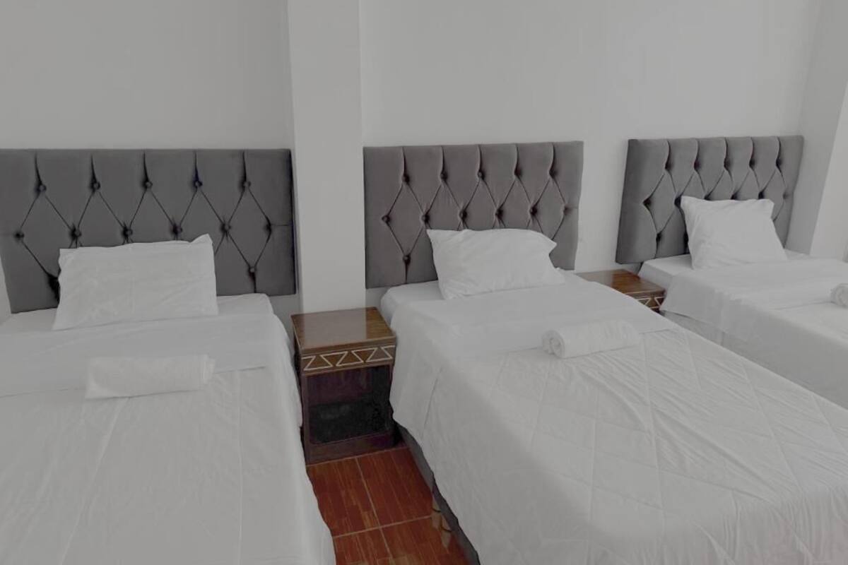 Habitación de hotel con tres camas, cada una con cabecero gris capitonado, ropa de cama blanca y una mesita de noche de madera entre las camas.