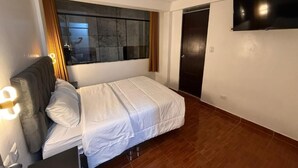 Een slaapkamer met een bed, een aan de muur bevestigde televisie, een raam met gordijnen en een deur.