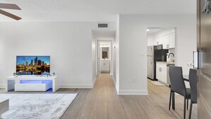 Un apartamento moderno con sala de estar que incluye un televisor de pantalla plana, comedor con mesa y sillas, y cocina con gabinetes blancos y refrigerador.