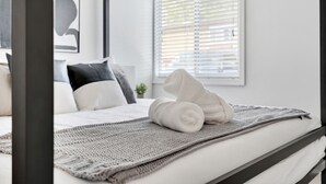 Una cama bien hecha con una manta blanca y toallas enrolladas, un plaid de punto gris y una ventana con persianas.