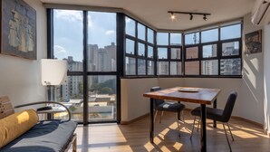 A modern room with a large window, a wooden table, two chairs, a sofa, and a lamp. - Apto em Campo Belo 20min Aeroporto CGH (São Paulo)