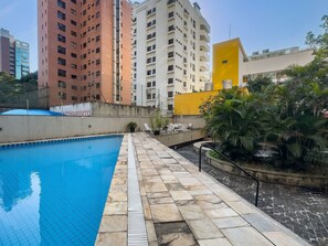 Piscina com ladrilhos de pedra, rodeada por uma vedação, e adjacente a um edifício com varandas.