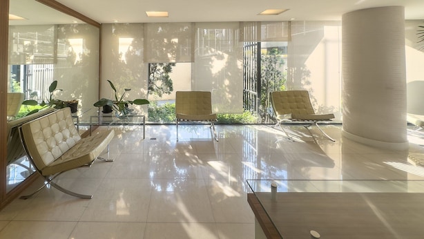 A modern lobby with a glass partition, two cushioned chairs, a large column, and a view of greenery outside. - Apto em Campo Belo 20min Aeroporto CGH (São Paulo)