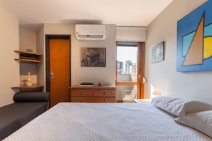 A bedroom with a bed, a dresser, a desk, and a chair. There is an air conditioning unit on the wall, a painting, and a window with a view of buildings. - Apto em Campo Belo 20min Aeroporto CGH (São Paulo)