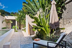Un área junto a la piscina con una silla de descanso, una mesa con una toalla y grandes plantas de plátano.