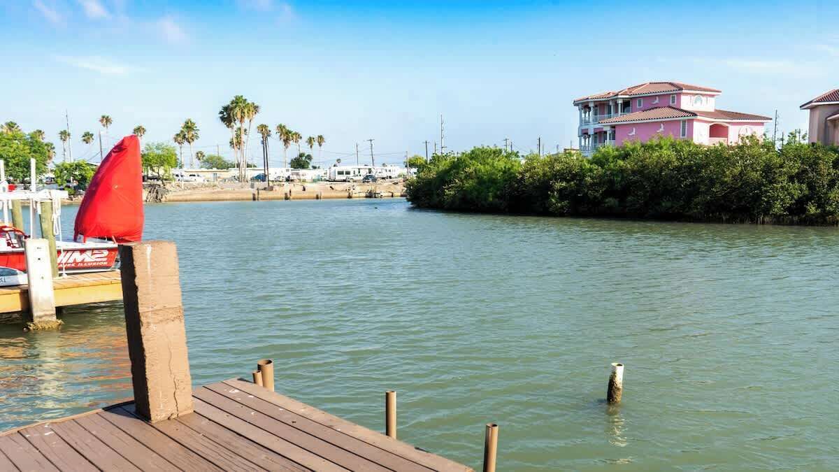 A wooden dock extending into calm water with a red canopy, a pink building, and palm trees in the background.