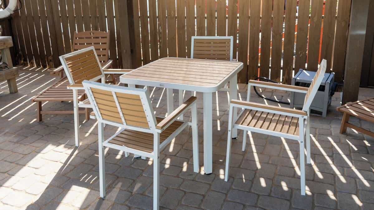 Un patio exterior con una mesa de madera y cuatro sillas, una mesa pequeña con una silla, y un banco de madera.