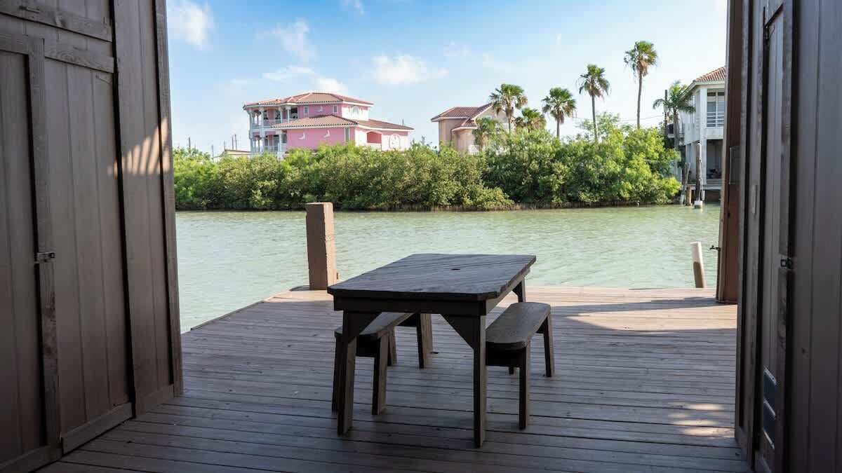 A wooden deck with a table and bench, overlooking calm water and buildings in the background.