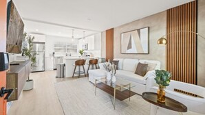 A modern living room with a white sofa, a glass coffee table, and a kitchen in the background. - Central Cozy Home | Free Crib & Parking Space (Tampa)