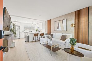 Ein modernes Wohnzimmer mit einer Couch, einem Couchtisch und Barhocker. Im Hintergrund ist eine Küche mit Kühlschrank und Essbereich zu sehen.