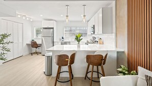 A modern kitchen with white cabinetry, a central island with stools, pendant lighting, and a view of the adjacent living area. - Central Cozy Home | Free Crib & Parking Space (Tampa)