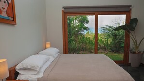 A bedroom with a bed, bedside lamps, a window with a view of greenery, and a painting on the wall. - Ocean View Studio at Taru Rentals (Montezuma)