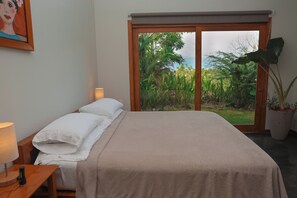 A bedroom with a bed, bedside lamps, a window with a view of greenery, and a painting on the wall. - Ocean View Studio at Taru Rentals (Montezuma)