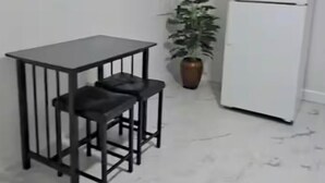 Ett litet kök med ett vitt kylskåp, ett svart metallbord med två pallar och en krukväxt.