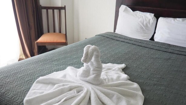 Una cama con una toalla blanca doblada en forma de cisne, una silla de madera y un cabecero oscuro.