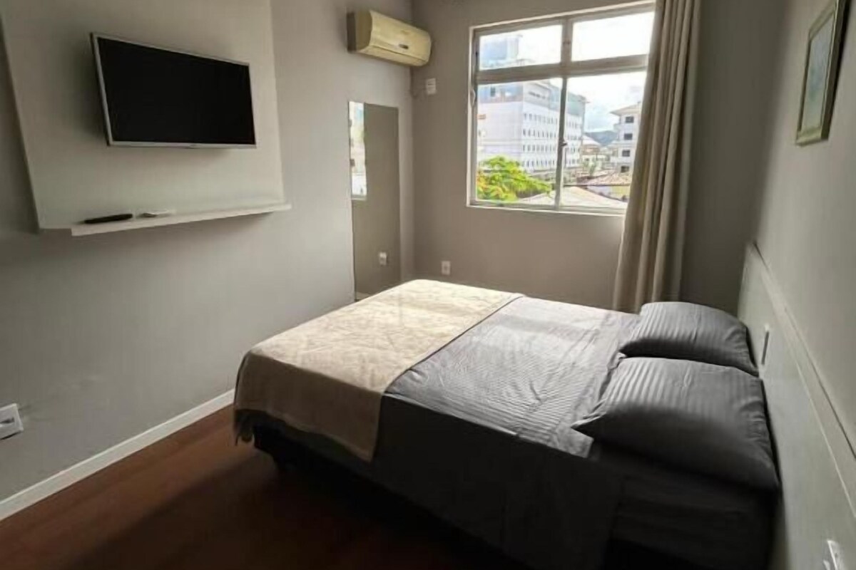 Una camera da letto con un letto, una TV a muro, un condizionatore d'aria e una finestra con vista sugli edifici.