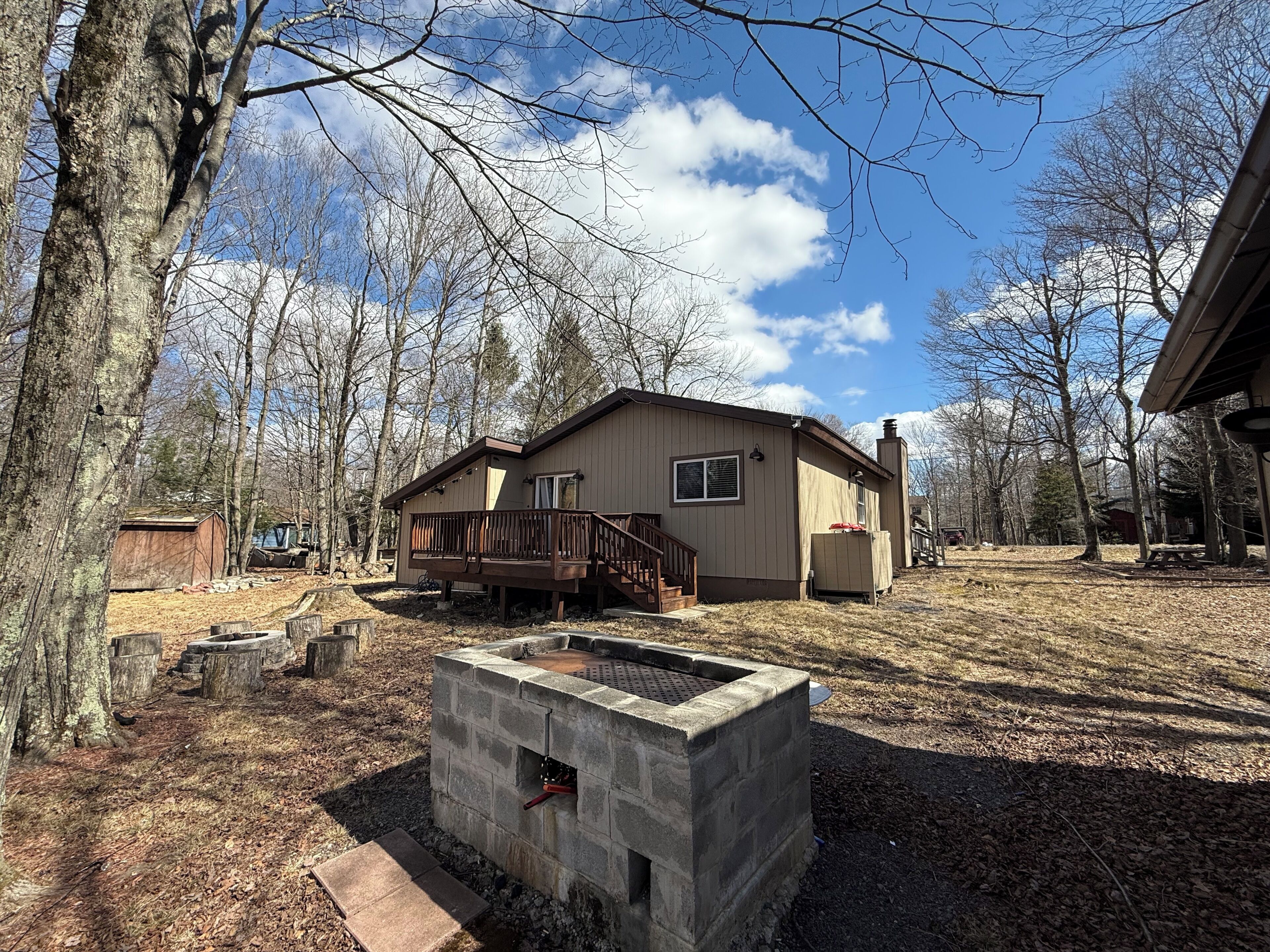 A single-story house with a deck, a stone fire pit, and a wooded backyard.
