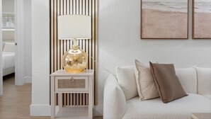 Een moderne woonkamer met een witte bank, een lamp op een houten nachtkastje en een schilderij met een strandthema aan de muur.