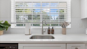 Een moderne keuken met een roestvrijstalen spoelbak, witte kasten, een raam met zonwering en een potplant.