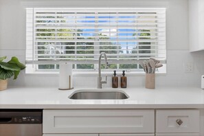 Eine moderne Küche mit Spüle aus Edelstahl, weißen Schränken, einem Fenster mit Jalousien und einem Blumentopf.