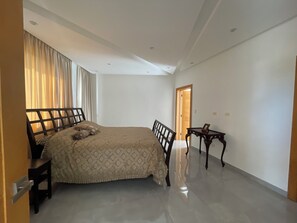 Quarto com cama, cadeira, mesa pequena e porta que dá para outro cômodo.