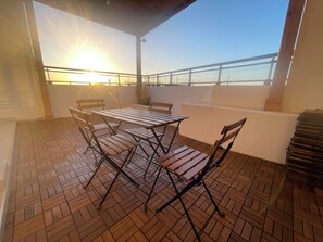 Um pátio externo com mesa e cadeiras, corrimão e vista para o mar.