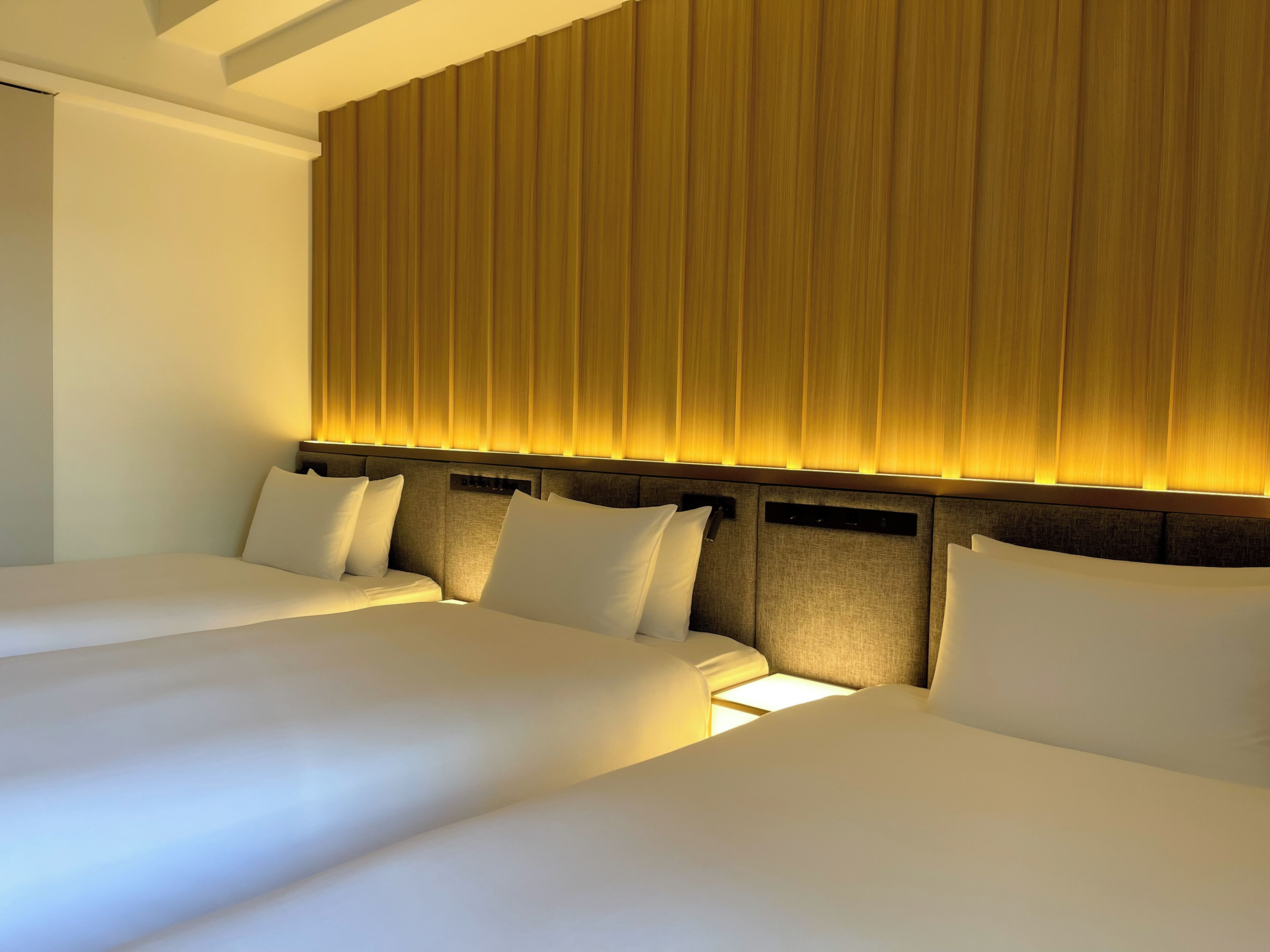 A hotel room with two beds, a headboard with vertical lines, and a light source behind the headboard.