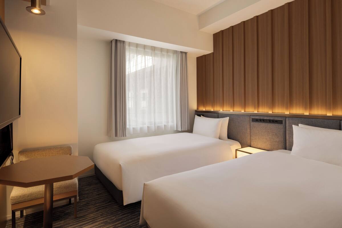 A hotel room with two beds, a desk, a TV, and a large window with curtains.