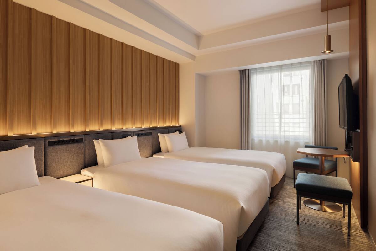 A hotel room with three beds, a small table, a TV, and a large window with curtains.