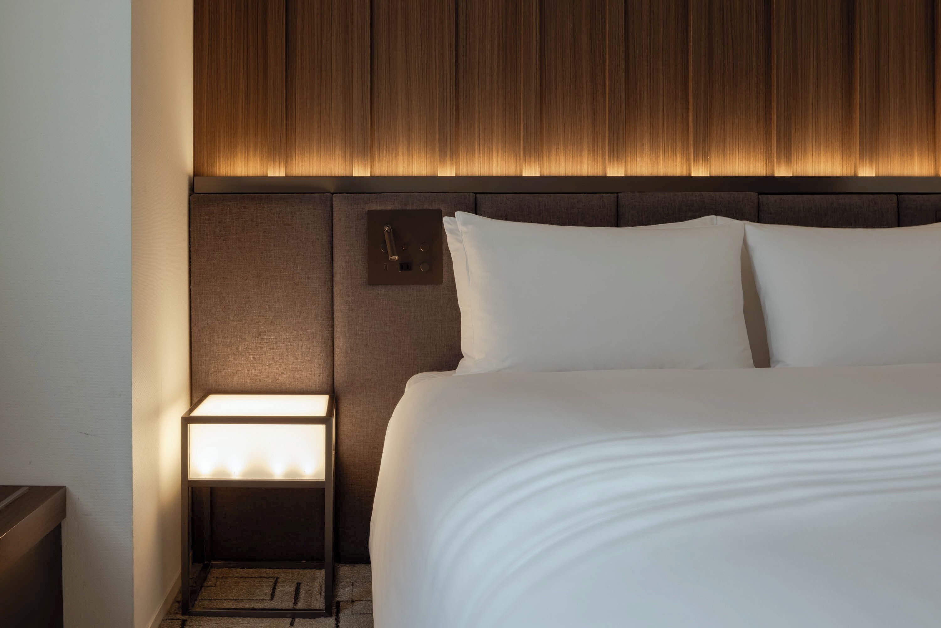 Una habitación de hotel con una cama grande, una mesita de noche con una lámpara y un cabecero de madera.