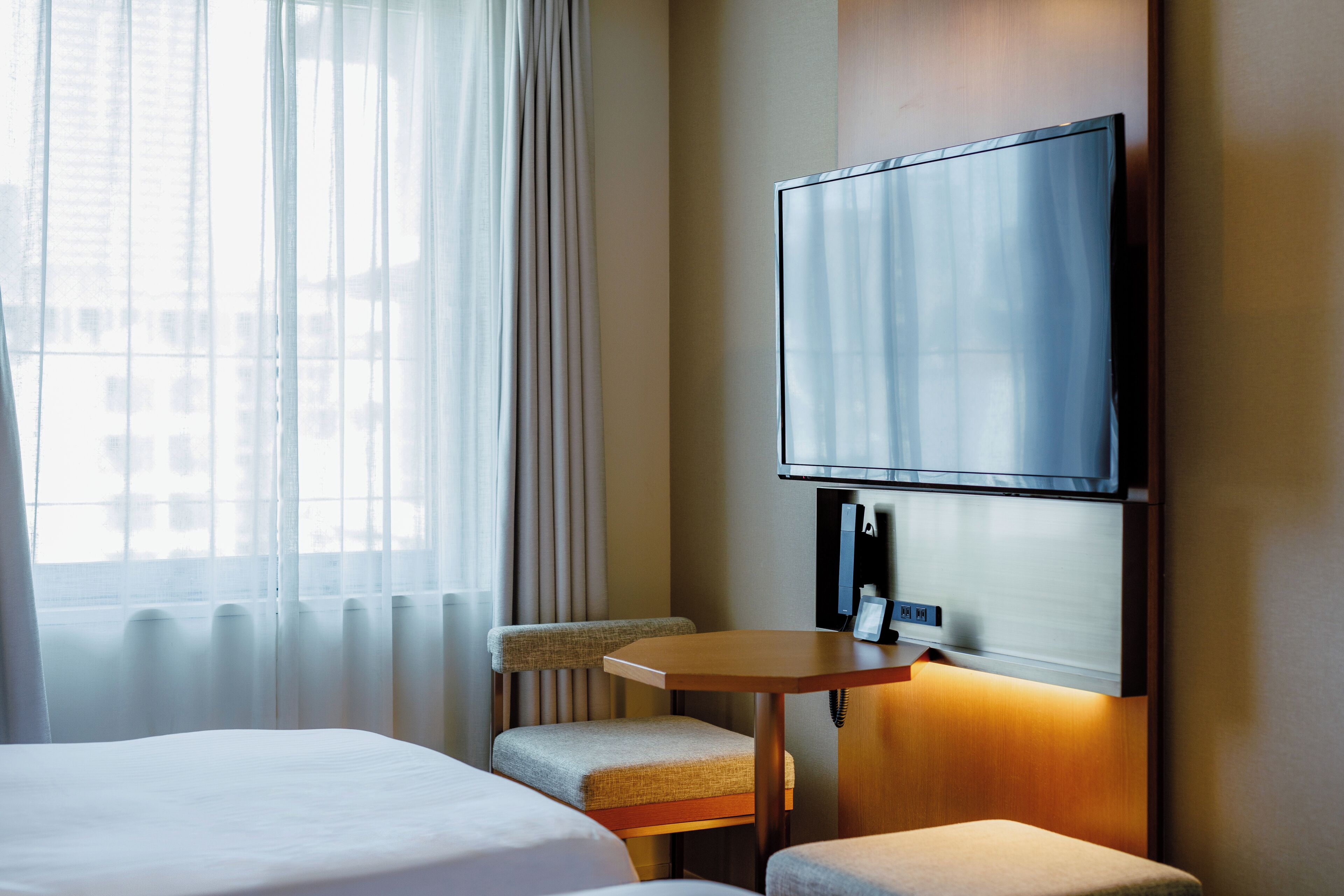 Ein Hotelzimmer mit Bett, Sessel, kleinem Tisch, Wandfernseher und einem Fenster mit Vorhängen.
