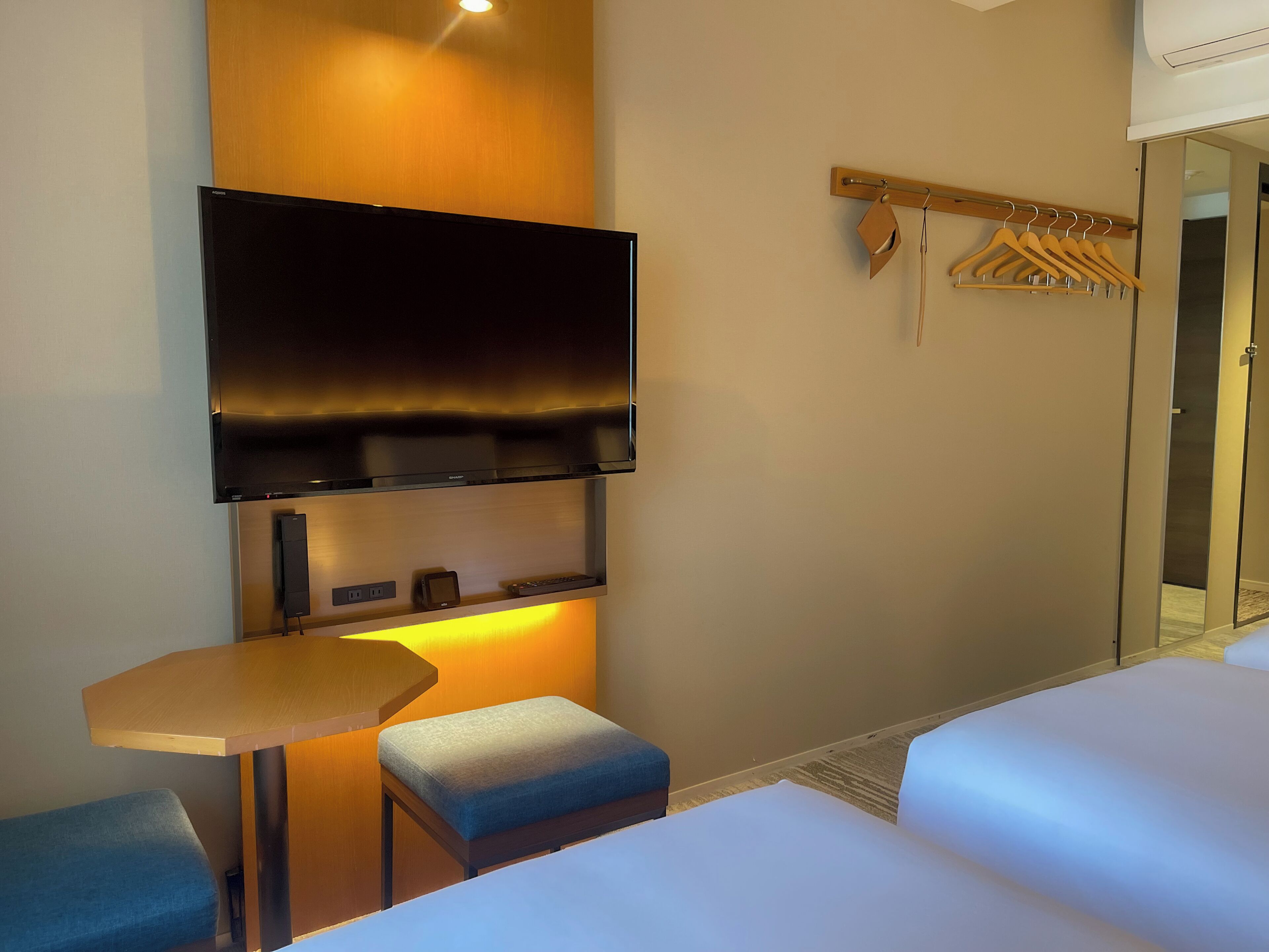 Una habitación de hotel con una televisión montada en la pared, una mesita, un banco y una cama.