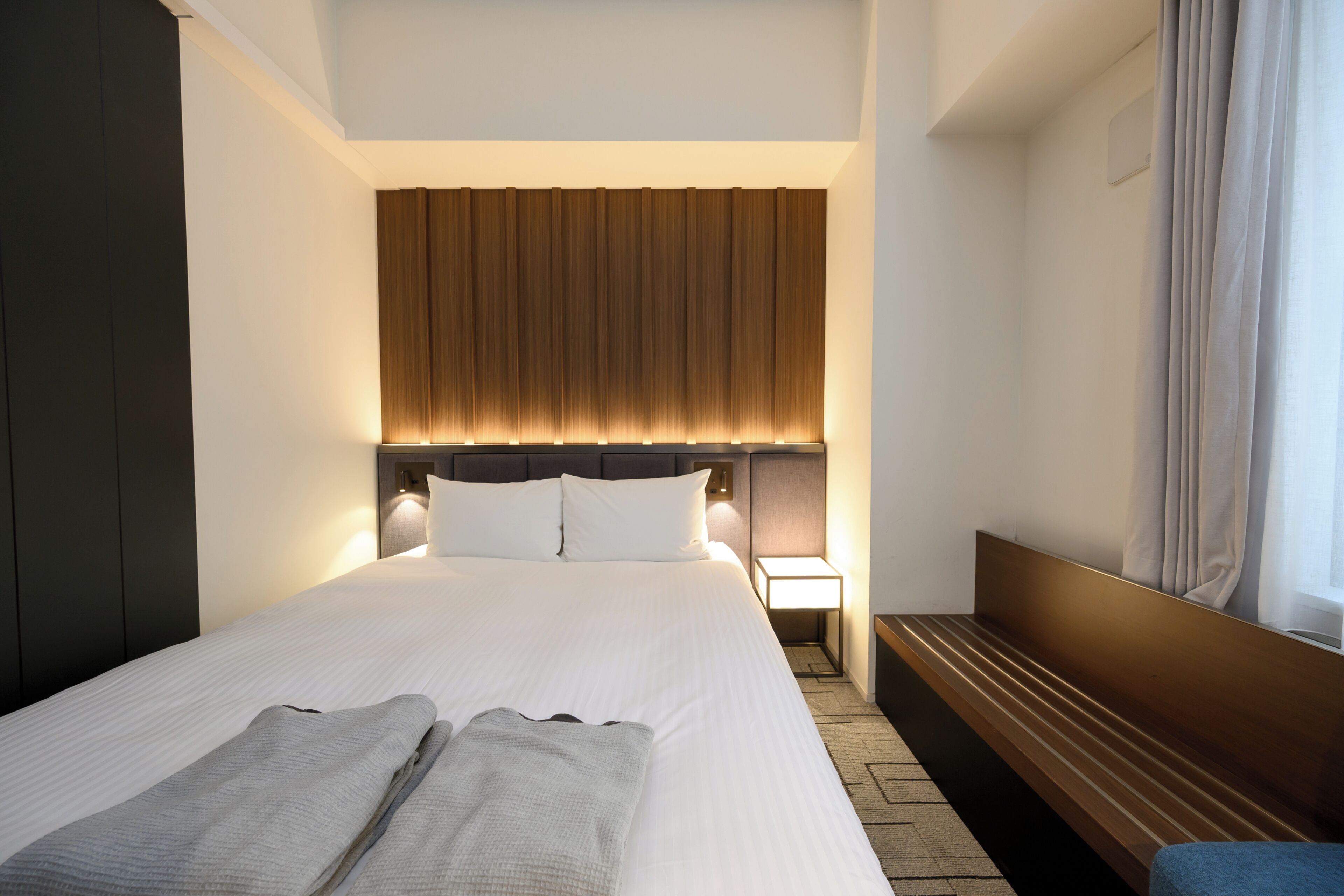 Una habitación de hotel moderna con una cama grande, una mesita de noche y un ventanal con cortinas.