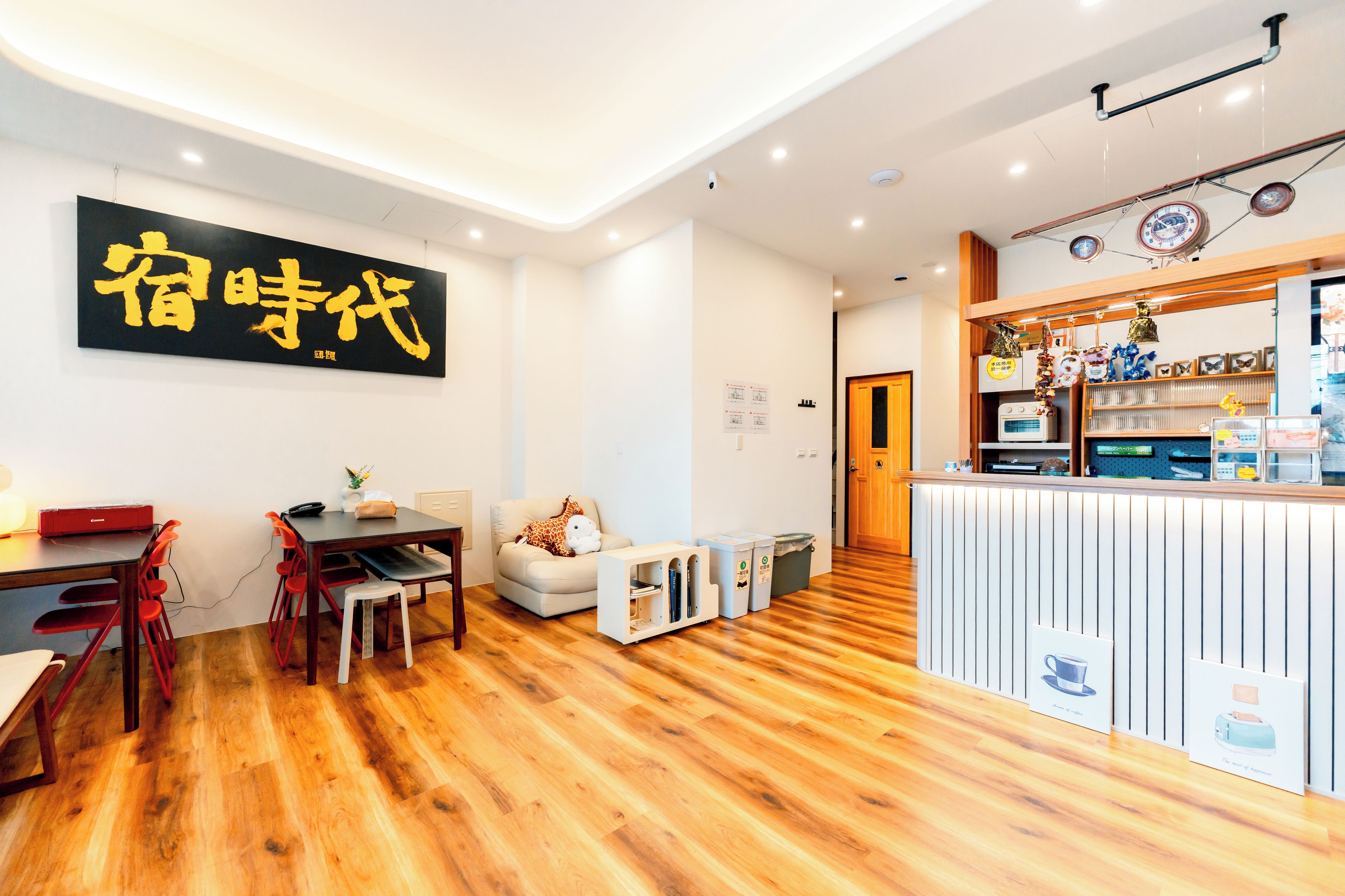 A modern interior with a wooden floor, a dining area with a table and chairs, a sofa with a giraffe toy, and a reception desk with various items.