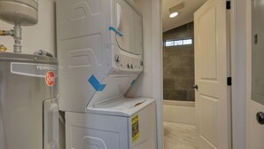 Ein Waschraum mit einer weißen Waschmaschine und einem Trockner, einem Warmwasserbereiter und einem Duschbereich, der durch eine Glastür sichtbar ist.