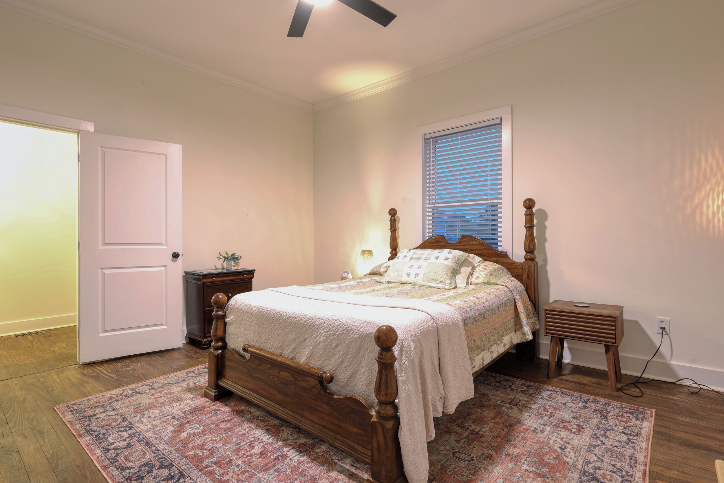A bedroom with a wooden bed, a nightstand, a bedside table, a window with blinds, and a door.