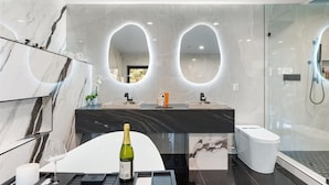 Un baño moderno con tocador de mármol, dos lavamanos, cabina de ducha de cristal y una botella de champán en la encimera.