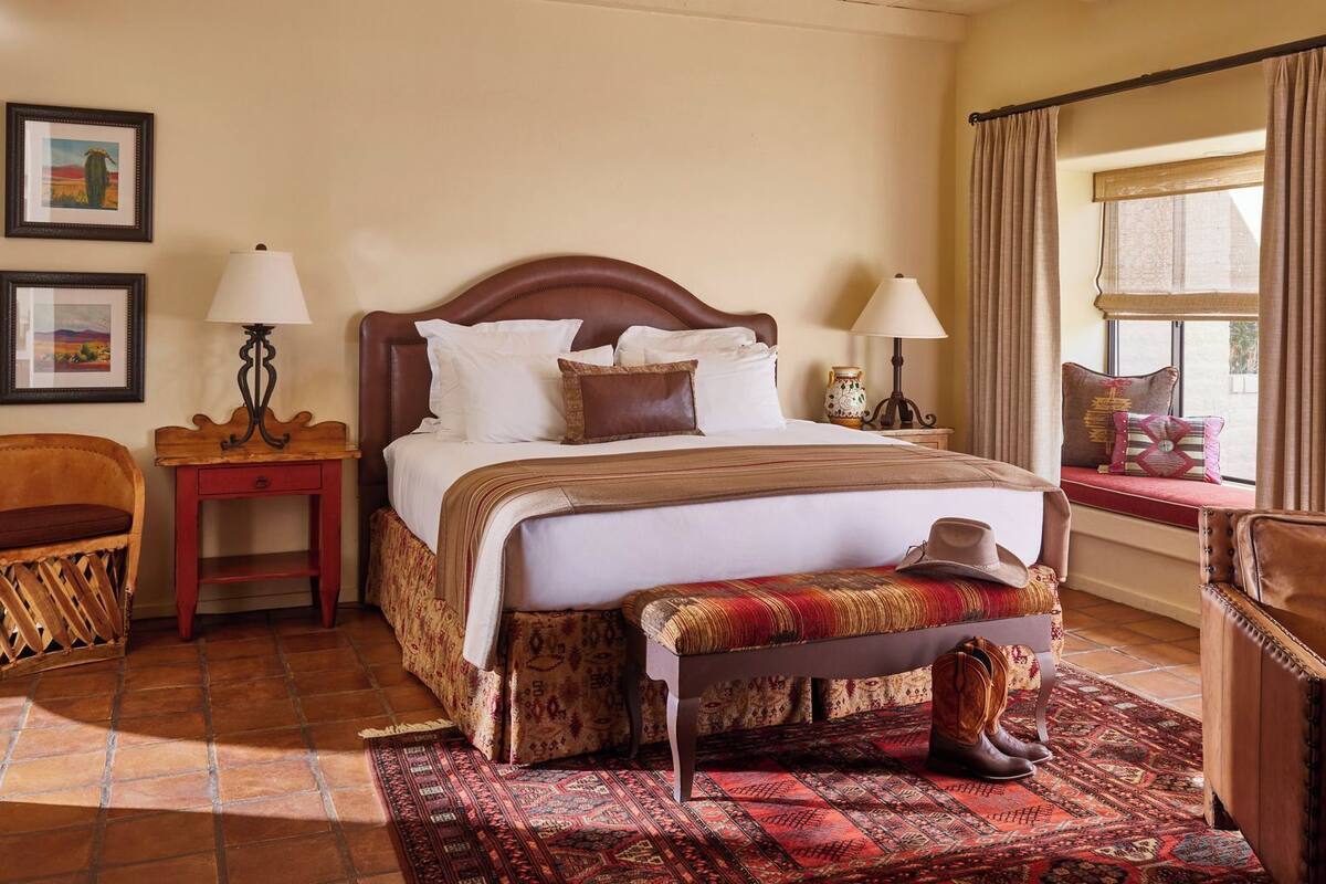 Un cuarto de hotel ordenado con una cama, mesitas de noche, un banco, una silla y una ventana con cortinas.